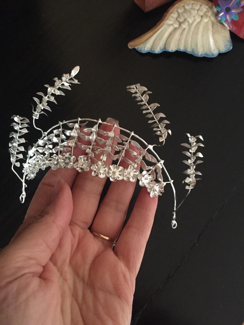 Puede incluir: Corona de plata con dise&ntilde;o floral. La corona est&aacute; hecha de alambre y tiene peque&ntilde;as flores y hojas de plata.