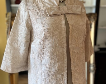 May include: A long, pale pink, brocade jacket with a large bow at the collar. The jacket has a textured, floral pattern and a front closure.
