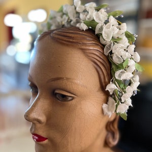 May include: A white flower crown with green leaves on a mannequin head. The crown is made of fabric flowers and is a vintage style.