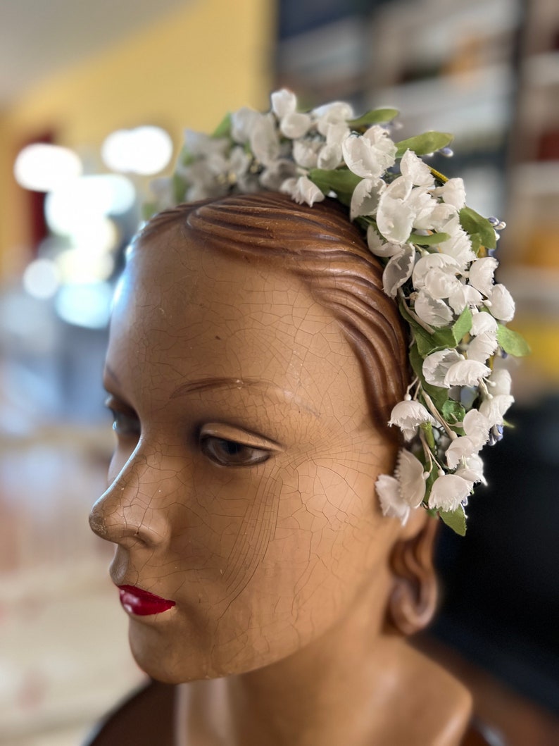 May include: A white flower crown with green leaves on a mannequin head. The crown is made of fabric flowers and is a vintage style.