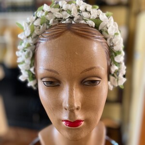 May include: A white flower crown with green leaves, worn on a mannequin head. The crown is made of artificial flowers and is a vintage style.