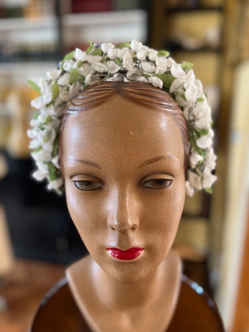 May include: A white flower crown with green leaves, worn on a mannequin head. The crown is made of artificial flowers and is a vintage style.
