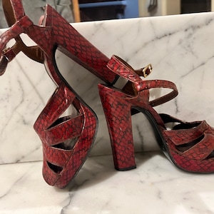 May include: Red high-heeled sandals with a snakeskin pattern. The open-toe shoes have thick heels, ankle straps, and a platform sole. The sandals are made of a textured material and feature a buckle closure.