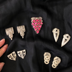 May include: A set of eight vintage silver tone brooches with rhinestones and pink glass stones. The brooches are in the shape of grapes, leaves, and geometric shapes.