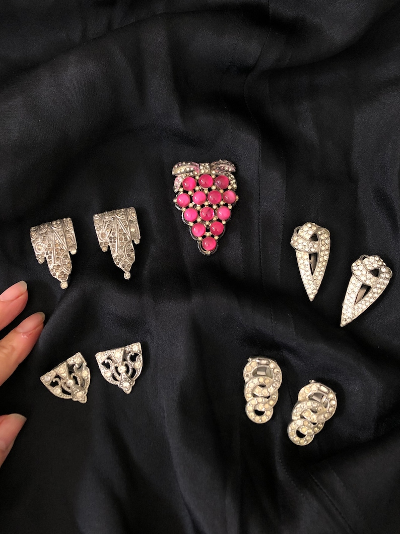 May include: A set of eight vintage silver tone brooches with rhinestones and pink glass stones. The brooches are in the shape of grapes, leaves, and geometric shapes.