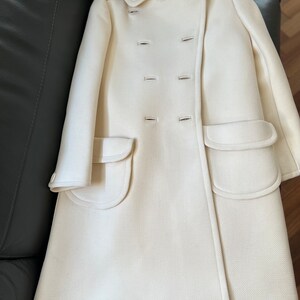 May include: A white double-breasted coat with two large pockets on the front. The coat has a collar and is made of a soft, textured fabric.