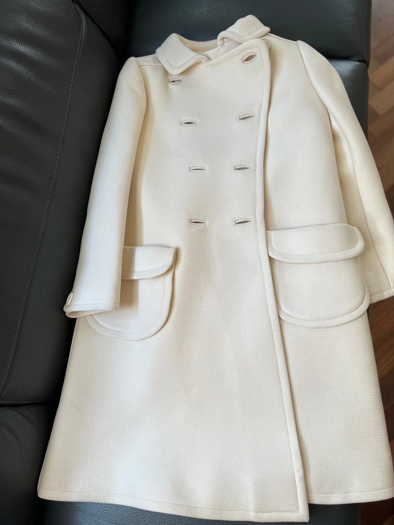 May include: A white double-breasted coat with two large pockets on the front. The coat has a collar and is made of a soft, textured fabric.