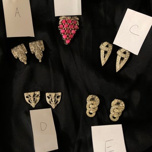 May include: Five pairs of vintage rhinestone jewelry pieces labeled A through E.  Piece A is a pair of silver tone earrings with a floral design. Piece B is a pink rhinestone grape cluster brooch. Piece C is a pair of silver tone earrings with a pointed design. Piece D is a pair of silver tone earrings with a geometric design. Piece E is a pair of silver tone earrings with a circular design.