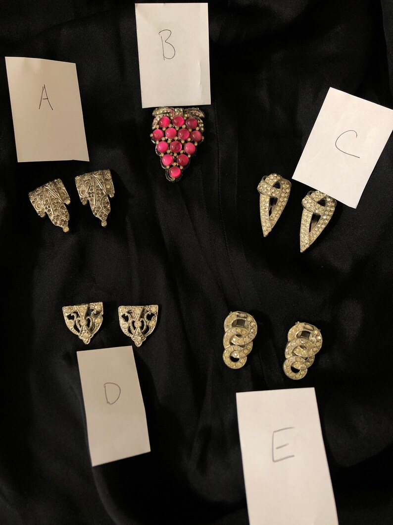 May include: Five pairs of vintage rhinestone jewelry pieces labeled A through E.  Piece A is a pair of silver tone earrings with a floral design. Piece B is a pink rhinestone grape cluster brooch. Piece C is a pair of silver tone earrings with a pointed design. Piece D is a pair of silver tone earrings with a geometric design. Piece E is a pair of silver tone earrings with a circular design.