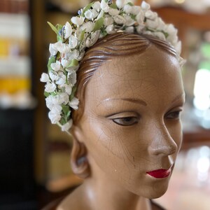 May include: A white flower crown with green leaves and silver accents. The crown is worn on a wooden mannequin head.