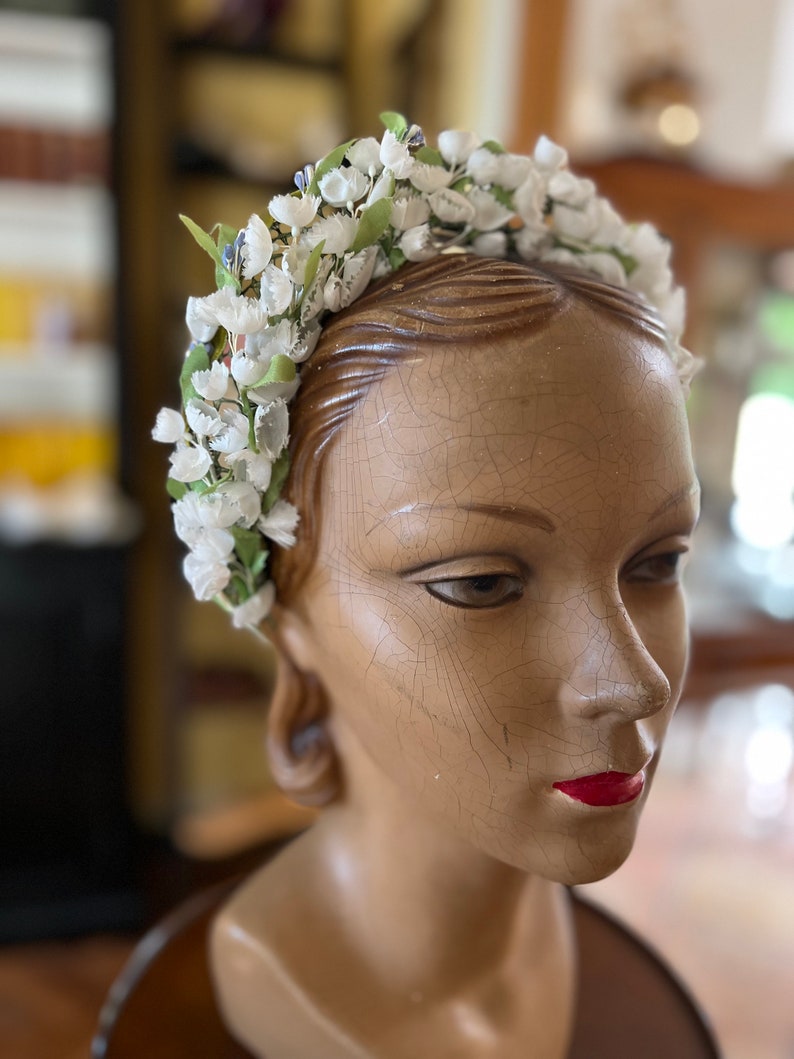 May include: A white flower crown with green leaves and silver accents. The crown is worn on a wooden mannequin head.