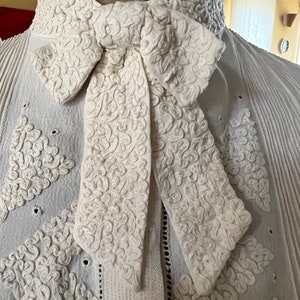 Op de afbeelding: Een wit katoenen blouse met een hoge kraag en een grote, ingewikkelde strik. De strik is gemaakt van dezelfde stof als de blouse en heeft een delicaat, bloemig patroon.