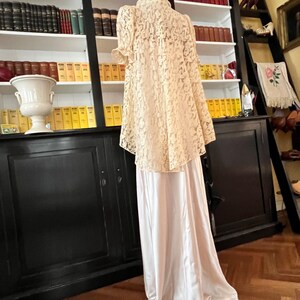 Puede incluir: Una blusa de encaje blanco con cuello alto y mangas cortas, combinada con una falda de sat&eacute;n blanco. El atuendo se muestra en un maniqu&iacute; frente a una estanter&iacute;a.