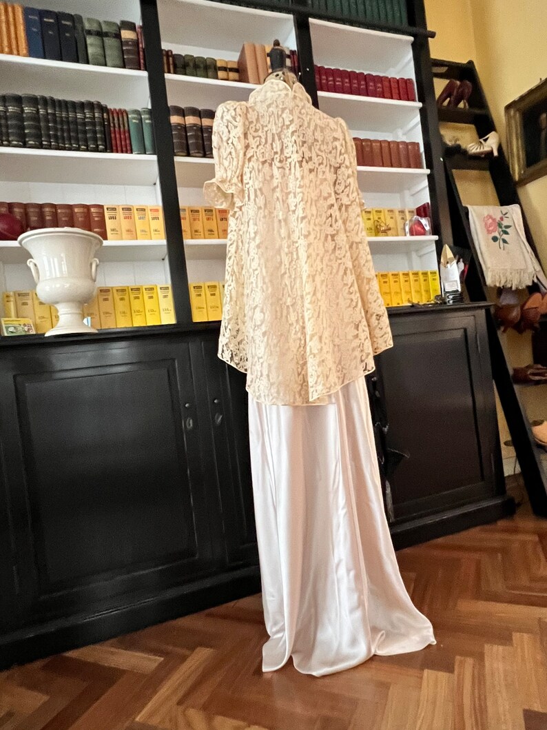 Puede incluir: Una blusa de encaje blanco con cuello alto y mangas cortas, combinada con una falda de sat&eacute;n blanco. El atuendo se muestra en un maniqu&iacute; frente a una estanter&iacute;a.