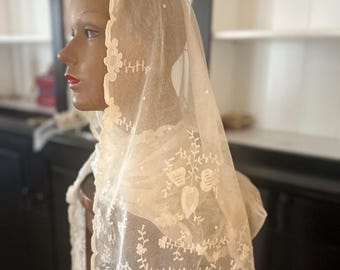 May include: A cream-coloured, sheer lace veil with floral and leaf embroidery. The veil is draped over a mannequin head, showcasing the delicate details of the lace. The edges are scalloped, and the design includes small, scattered dots.