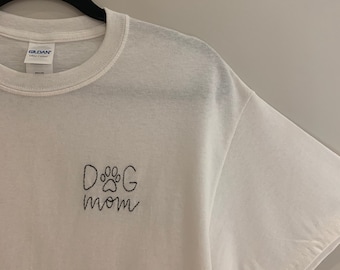 May include: White t-shirt with black embroidered text that says "DOG mom" with a paw print in the "O".
