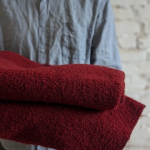 May include: A folded, deep red blanket with a textured, nubby surface. The blanket is held by a person wearing a light blue button-down shirt. The background is a white brick wall.