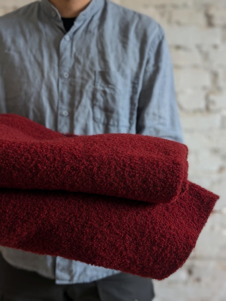 May include: A folded, deep red blanket with a textured, nubby surface. The blanket is held by a person wearing a light blue button-down shirt. The background is a white brick wall.