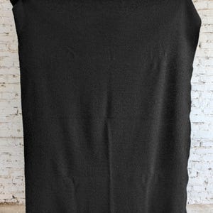 May include: A large, solid black blanket is held up against a white brick wall. The blanket has a textured surface, and is being held up by two hands, with the brick wall in the background.