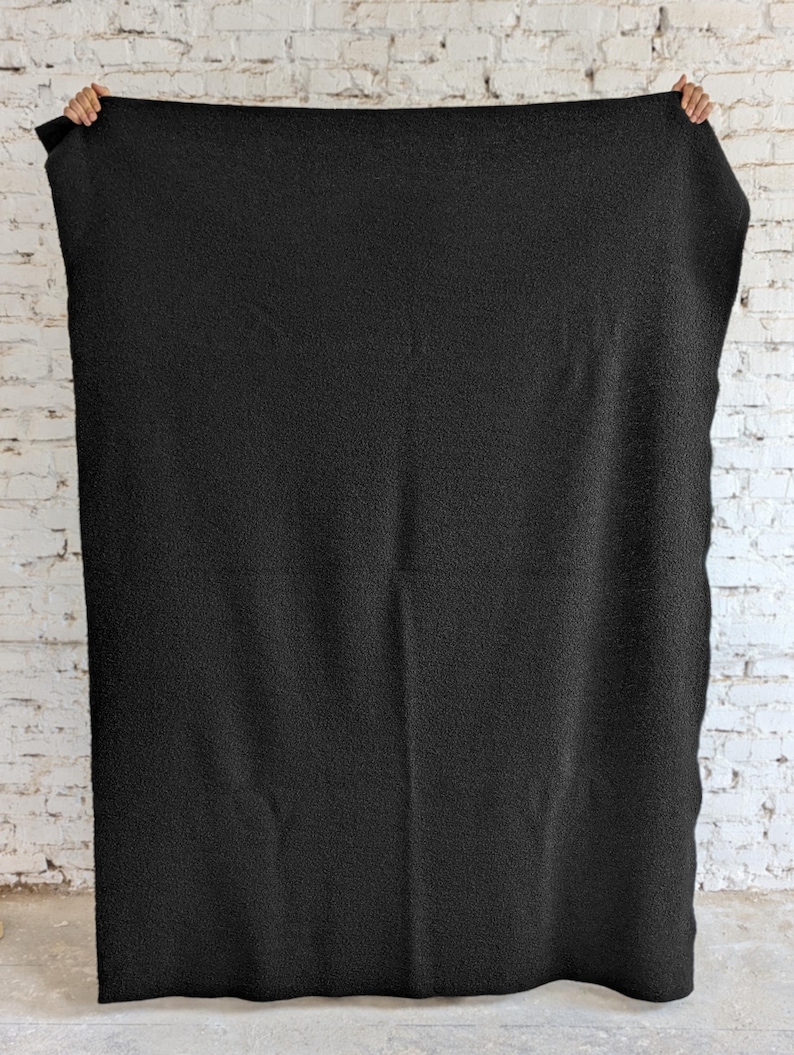 May include: A large, solid black blanket is held up against a white brick wall. The blanket has a textured surface, and is being held up by two hands, with the brick wall in the background.