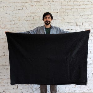 May include: A large, black, plush blanket is held up in front of a white brick wall. The blanket is rectangular and appears soft. The person holding the blanket is wearing a gray shirt and dark pants.