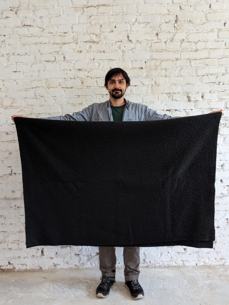 May include: A large, black, plush blanket is held up in front of a white brick wall. The blanket is rectangular and appears soft. The person holding the blanket is wearing a gray shirt and dark pants.