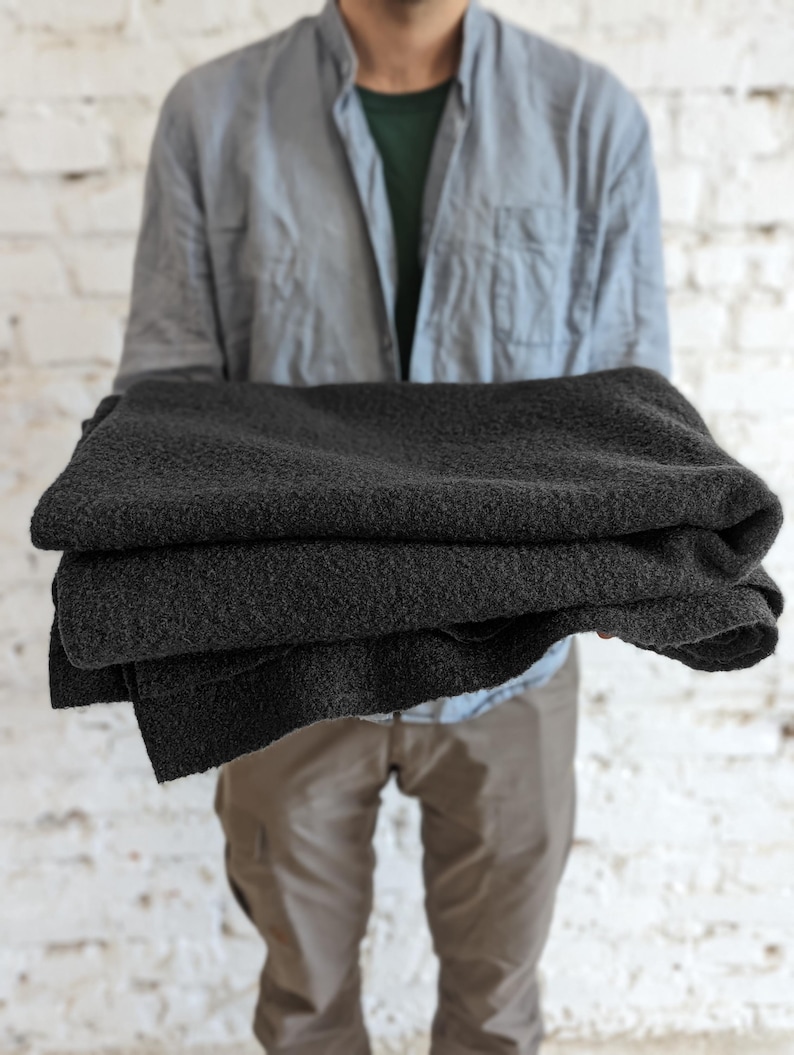 May include: A stack of folded, dark gray blankets. The blankets have a textured surface and appear soft. The person holding the blankets is wearing a light blue button-down shirt and khaki pants, standing in front of a white brick wall.