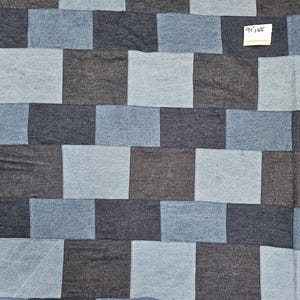 May include: A fabric featuring a repeating checkerboard pattern of alternating dark charcoal gray and light blue rectangular patches. A small white rectangular label is attached to the upper right corner with the text 94" x WOF written in black ink.