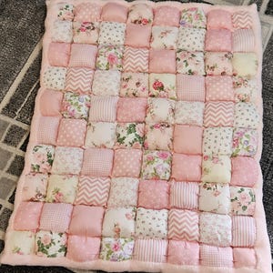Puede incluir: Una colcha rectangular tipo puff construida con numerosos cuadrados de tela pequeños y acolchados en varios tonos de rosa, blanco y crema. Los cuadrados individuales presentan diversos patrones, incluyendo estampados florales, lunares, rayas y chevrones.