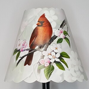 May include: A white lampshade with a scalloped edge features a red cardinal perched on a branch with white and pink blossoms. The background is a light gray with a subtle floral pattern.