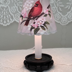 May include: A small table lamp with a white candle and a gray lampshade featuring a red cardinal perched on a branch with pink blossoms. The lampshade has a scalloped edge.