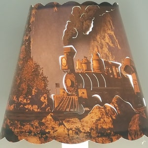 May include: A brown lampshade with a scalloped edge features a cutout design of a vintage steam locomotive with smoke billowing from the chimney. The locomotive is set against a backdrop of a mountain range and trees.