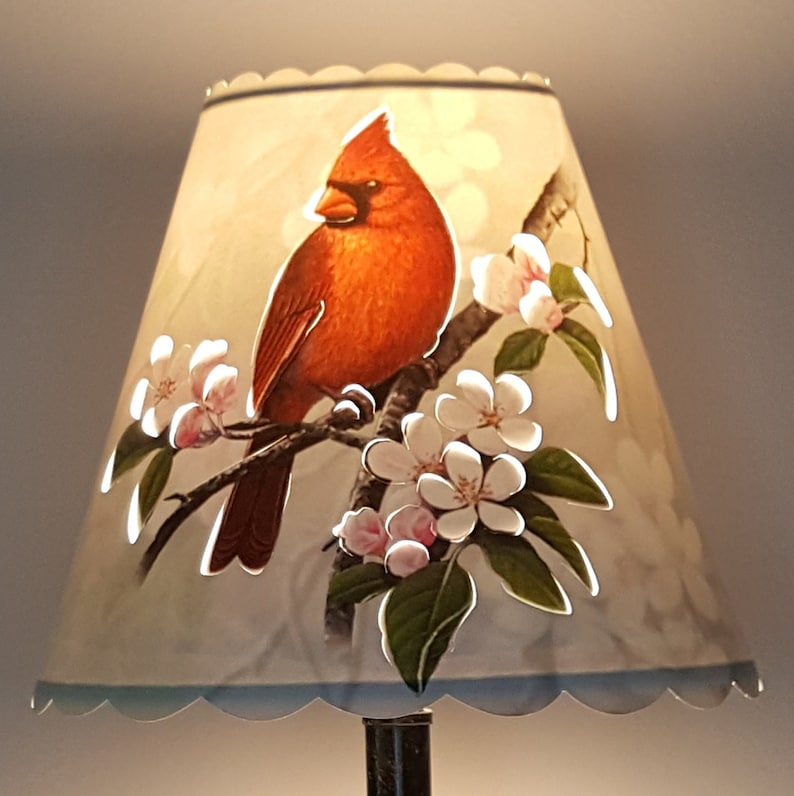 May include: A white lampshade with a scalloped edge features a red cardinal perched on a branch with white and pink blossoms. The lampshade has a light blue trim.