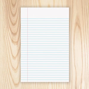 May include: A white, lined notepad with a red margin line and blue horizontal lines. The notepad is rectangular and rests on a light-colored wooden surface. The lines are evenly spaced, creating a grid pattern.