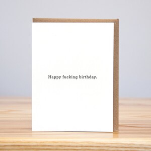 May include: A white card with brown edges. The card has the text "Happy fucking birthday." printed in black.