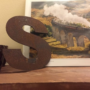 Op de afbeelding: Een roestige metalen letter S, staand op een houten oppervlak, voor een ansichtkaart met een trein op een brug.
