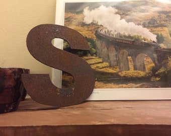 Può includere: Una lettera S in metallo arrugginito, in piedi su una superficie di legno, davanti a una cartolina che mostra un treno su un ponte.