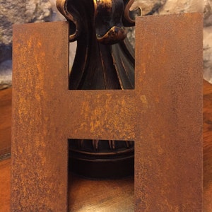 Op de afbeelding: Een roestige metalen letter H, staand op een houten oppervlak. De letter is donkerbruin van kleur met een ruw, getextureerd oppervlak.