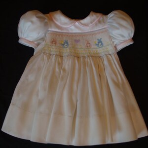 May include: A white smocked dress with a pink collar and puffed sleeves. The dress has a floral embroidery design with birds and hearts.