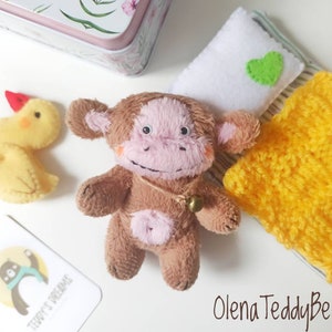 May include: A small brown plush monkey with a light pink face and belly, wearing a tiny gold bell on a string. Beside it are a yellow felt chick, a white felt pillow with a green heart, a yellow knitted blanket, and a tag labeled Teddy's Dreams.