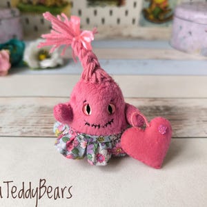 Può includere: Una creatura di peluche rosa fatta a mano con una bocca cucita sorridente e occhi a bottone. Indossa una gonna con stampa floreale e tiene in mano un cuore rosa. La creatura ha una testa a ciuffi con un fiocco rosa. Il testo "Olena TeddyBears" è in basso.
