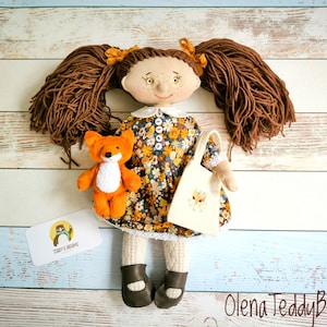 May include: A handmade doll with brown yarn hair, freckles, and a floral dress. It holds a small orange teddy bear and a tote bag with a fox design. The doll wears brown shoes and has a white collar. The doll is a handmade item.