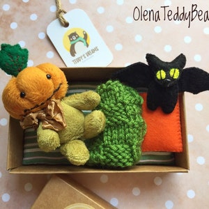 May include: A small, handmade pumpkin-shaped plush toy with a green stem and a bow. The toy is sitting in a brown box with a green knitted blanket, an orange felt cushion, and a black felt bat with yellow eyes. The box has a label that says "Teddy's Dreams".