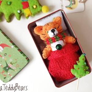 May include: A small plush tiger toy with orange and black stripes, wearing a green and red scarf and a green bell, tucked into a red knitted sleeping bag inside a rectangular metal tin. A small green felt tree is placed next to the tiger.
