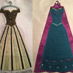 May include: Two embroidered felt dresses on hangers. The dress on the left is green and gold with black stripes. The dress on the right is teal with a purple cape and gold trim.