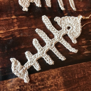 May include: A pair of crocheted fish bone earrings in a cream color. The earrings are made of yarn and have a delicate, intricate design.