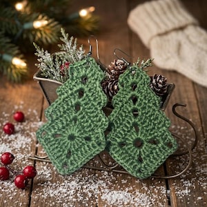 May include: A pair of green crochet earrings shaped like Christmas trees. Each earring features a tiered, openwork design with a textured yarn finish and a silver-toned metal hook for attachment. The earrings are displayed in a decorative metal sleigh.