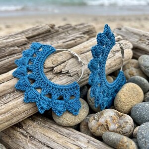 May include: A pair of hoop earrings featuring deep blue crochet thread wrapped around silver-toned metal hoops. The crochet work creates a scalloped, fan-like edge with textured, raised stitches along the outer perimeter of each circular earring.
