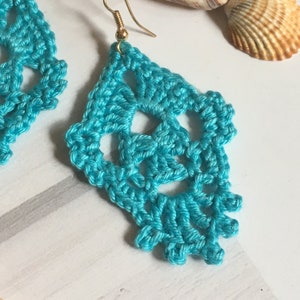 Op de afbeelding: Een paar turquoise gehaakte oorbellen met gouden haakjes. De oorbellen hebben de vorm van een traan en een delicaat, ingewikkeld ontwerp.