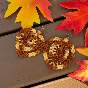May include: A pair of handmade crochet earrings shaped like leaves, featuring a spiral pattern in shades of brown and cream yarn. Each earring is accented with a small, light brown wooden bead and attached to a silver-toned metal fishhook ear wire.